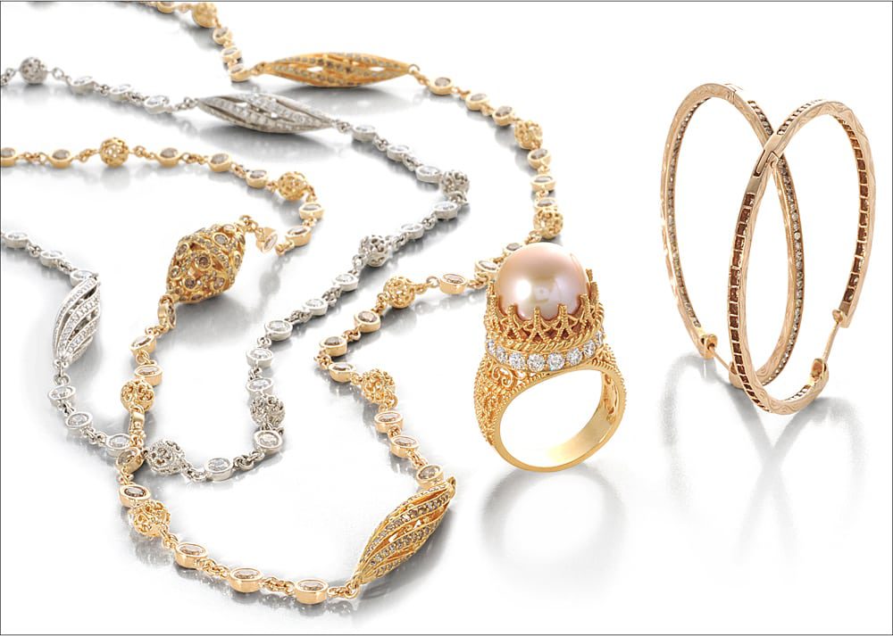 Ornate gold and silver jewelry collection on white background. Silver and diamond necklace, gold and diamond necklace, gold ring with diamond circling crown mount featuring large pink pearl and two gold bangles with brown diamonds.