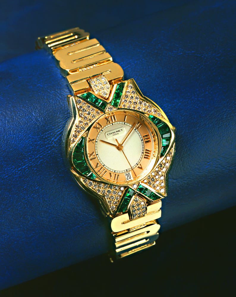 Fancy Chaumet men's watch with diamonds and emeralds and Roman Numerals on face. Band features stylized letters in gold all on blue vinyl background Product Photography