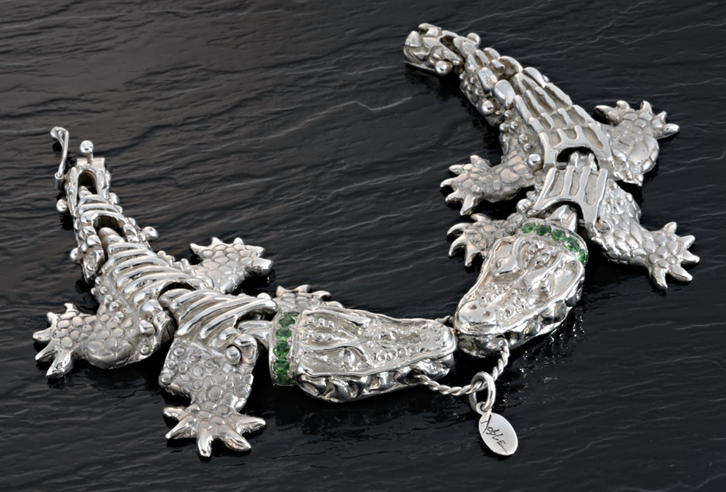 Silver Alligator bracelet with two alligators biting center ring. Alligators have collars with emeralds all on shiny black patterned background Product Photography