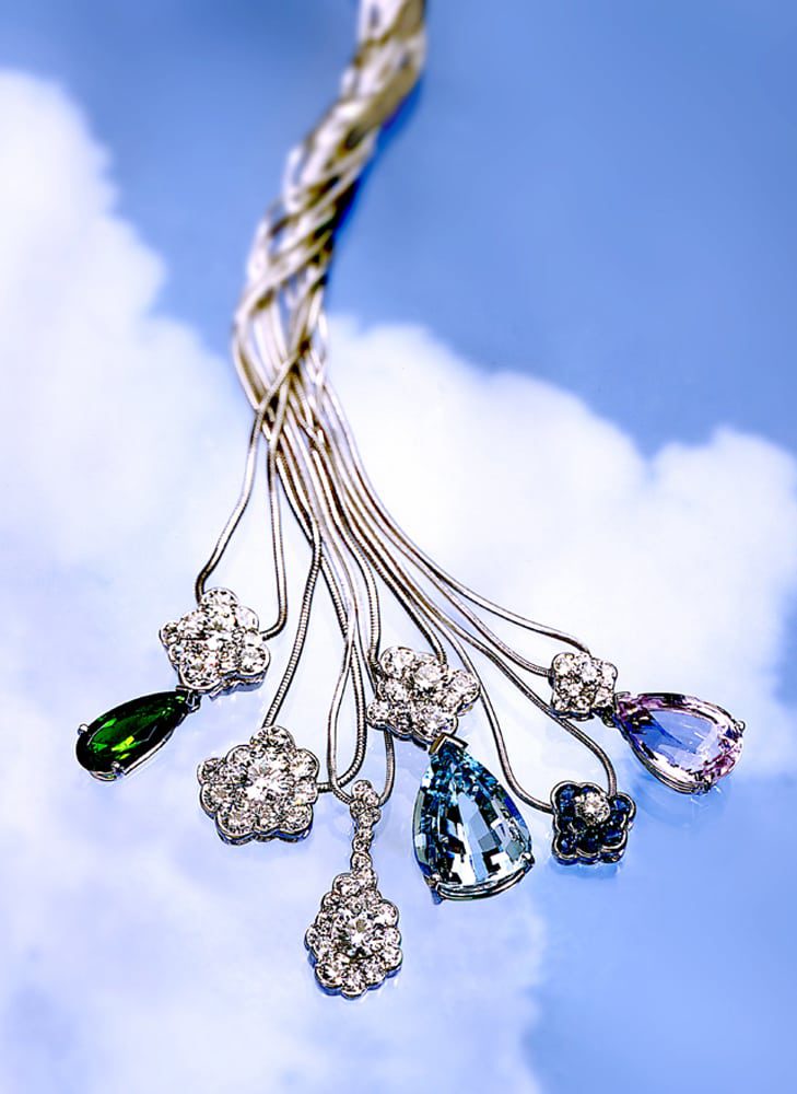 Six pendants on silver chains three with large colored stones and others with diamonds on blue sky and clouds background. Chains are all twisted up above the pendants.