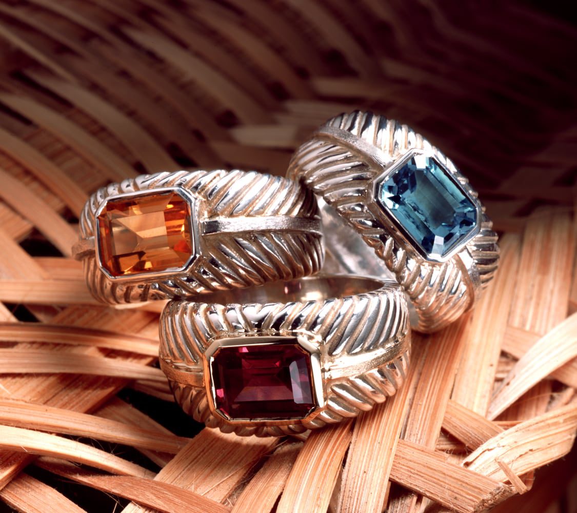 Three silver rings with a wicker pattern and yellow, blue and red stones, stacked on a wicker background